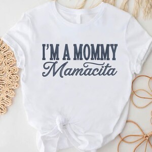 Op de afbeelding: Wit T-shirt met de marineblauwe tekst "I'M A MOMMY Mamacita". Het T-shirt is in de taille geknoopt. Decoratieve items zijn rond het T-shirt op een wit oppervlak geplaatst.