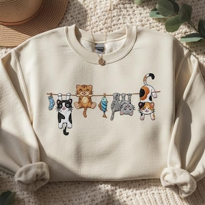 May include: Cream-coloured sweatshirt with an embroidered design of cats hanging from a washing line. The cats are in different colours and poses, with a fish and socks also on the line. A straw hat and necklace are in the background.