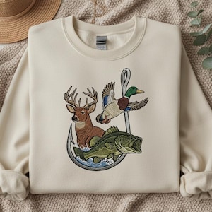 以下が含まれることがあります： 鹿、アヒル、魚が釣り針に描かれた刺繍デザインのクリーム色のスウェットシャツ。デザインには茶色、緑、青の色合いが使用されています。スウェットシャツは長袖で、クルーネックです。
