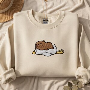 Peut inclure: Sweat-shirt crème avec un motif brodé d'un capybara brun reposant sur un canard blanc. Le canard a un bec et des pattes jaunes. Un chapeau de paille est visible en arrière-plan.