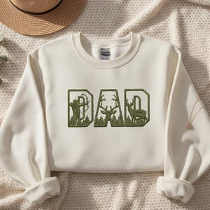 May include: Cream-colored sweatshirt with the word "DAD" embroidered in green. The letters feature outdoor-themed designs: an archer, a deer, and a deer silhouette. The sleeves are cuffed, and a straw hat is visible in the background.