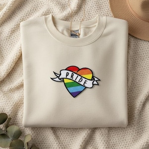 May include: A cream-coloured sweatshirt with a rainbow heart patch and the word "PRIDE" on a banner. The heart design features the colours of the rainbow. A gold necklace and a straw hat are visible in the background.