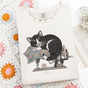 May include: Cream-colored t-shirt featuring an embroidered design of a sleeping black and white cat atop a vintage sewing machine. The cat rests on a floral pillow with a ball of yarn nearby. The shirt has the text "Comfort Colors" on the tag.