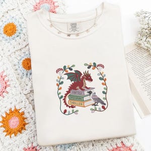 May include: A cream-colored t-shirt featuring an embroidered graphic of a red dragon perched on a stack of three books labeled Fantasy, Dystopian, and Thriller, accompanied by a black bird and a decorative vine border with small mushrooms.