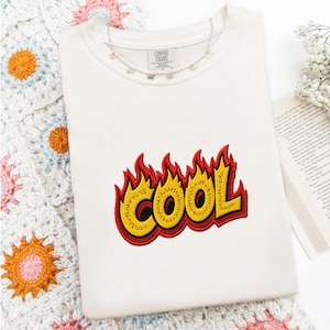 Op de afbeelding: Een crèmekleurig T-shirt met korte mouwen en een geborduurde afbeelding op de borst. Het ontwerp toont het woord COOL in dikke gele letters met een textuurpatroon, omlijnd in zwart en omringd door rode en oranje vlamvormen.