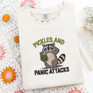 May include: A cream-colored t-shirt featuring an embroidered graphic of a raccoon holding a glass jar of green pickles. The text above the raccoon reads PICKLES AND in green, and the text below reads PANIC ATTACKS in black block lettering.