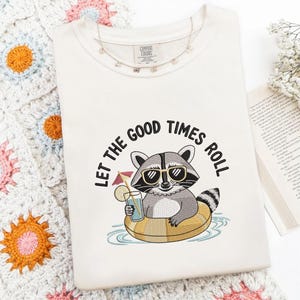 May include: Cream-colored t-shirt with the phrase "LET THE GOOD TIMES ROLL" encircling a raccoon wearing sunglasses and floating in a yellow inner tube. The raccoon is holding a drink with a lemon slice. A delicate necklace is draped on the shirt.