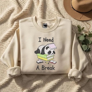 May include: Cream-coloured sweatshirt with the embroidered text "I Need A Break." The design features a sleeping panda on a stack of books. The panda is black and white, and the books are green and yellow.