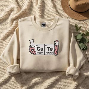 May include: Cream-coloured sweatshirt with a graphic design featuring the word "Cu Te" in a periodic table style, with hearts and laboratory glassware. The design includes the elements Copper and Tellurium.