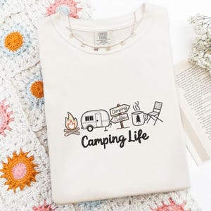 以下が含まれることがあります： 刺繍されたキャンプのデザインが施された、クリーム色のTシャツ。 デザインには、キャンプファイヤー、キャンピングカー、コーヒーカップ、椅子が含まれており、下に「Camping Life」の文字があります。 ネックレスが首元にあります。