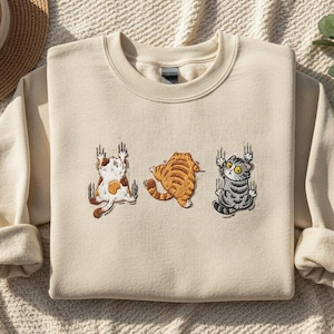 May include: Cream-colored sweatshirt featuring embroidered cartoon cats. The cats are in various poses, with one hanging on with its claws. The sweatshirt is made of a soft material and has a crew neck.