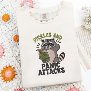 Puede incluir: Una camiseta de color crema con un gráfico bordado de un mapache que sostiene un frasco de vidrio lleno de pepinillos verdes. El texto sobre el mapache dice PICKLES AND en verde, y el texto debajo dice PANIC ATTACKS en color negro.