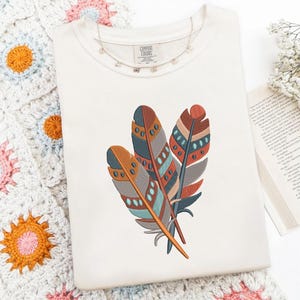 May include: Cream-colored t-shirt with an embroidered design of three colorful feathers. The feathers feature shades of orange, brown, teal, and blue, with intricate patterns. The shirt has the "Comfort Colors" label.