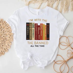 Può includere: T-shirt bianca con la scritta "I'M WITH THE BANNED ALL THE TIME" e una grafica di dorsi di libri. I dorsi dei libri sono di vari colori e includono titoli come "1984" e "Beloved".