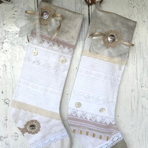 Può includere: Due calze di Natale in tessuto bianco e beige, decorate con pizzo, bottoni e fiocchi decorativi. Le calze hanno un aspetto vintage e artigianale, perfette per le decorazioni natalizie.