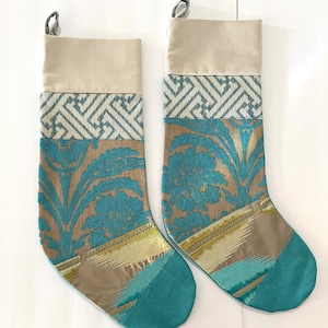Può includere: Due calze di Natale con la parte superiore beige, una fascia a motivi geometrici e un design floreale in verde acqua e oro. La punta della calza è verde acqua. Ogni calza ha un anello per appenderla.