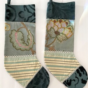 Può includere: Due calze natalizie decorative. Ogni calza presenta un motivo floreale nei toni del verde, rosa e oro. Le calze hanno una sezione a righe con bordi in pizzo e una punta in velluto verde scuro. C'è un anello nella parte superiore per appenderle.