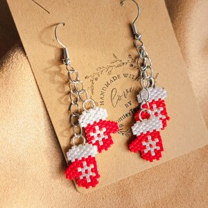 May include: A pair of handmade red and white beaded mitten earrings with silver-tone chain and hook closures. The mittens have a snowflake design. The earrings are displayed on a brown card with the text "Handmade with Love".