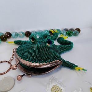 以下が含まれることがあります： 緑色のビーズで作られたカエルの形をした小銭入れ。黄色のアクセントとブロンズ色の留め金が付いています。カエルは大きな目を持ち、開いています。背景にはいくつかのビー玉があります。