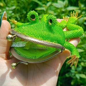 以下が含まれることがあります： 緑色のビーズで作られた手作りのカエル型の小銭入れ。カエルの目は金のアクセントで詳細に表現され、口を開くと小銭入れの中が見えます。小銭入れは、緑の葉を背景に、手で持たれています。
