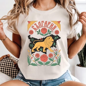 May include: Cream-colored t-shirt featuring a graphic of a lion, roses, and the word "България" in a decorative design. The lion is yellow, set against a black outline of a map. The roses are red.