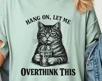 May include: A light green t-shirt with the text "Hang on, let me overthink this." The graphic features a detailed illustration of a cat holding a drink. The cat is gray and black with a serious expression.