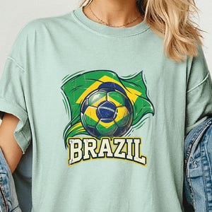 Pode incluir: Camiseta verde claro com uma bola de futebol e a bandeira brasileira. A palavra "BRAZIL" est&aacute; impressa em amarelo abaixo. A camiseta tem mangas curtas e um corte folgado.