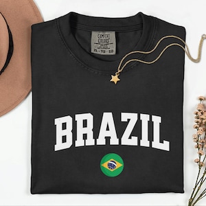 May include: Black t-shirt with the word "BRAZIL" in white block letters and a Brazilian flag graphic. A gold star pendant necklace rests on the shirt. The shirt is a size XL-TG-EG.