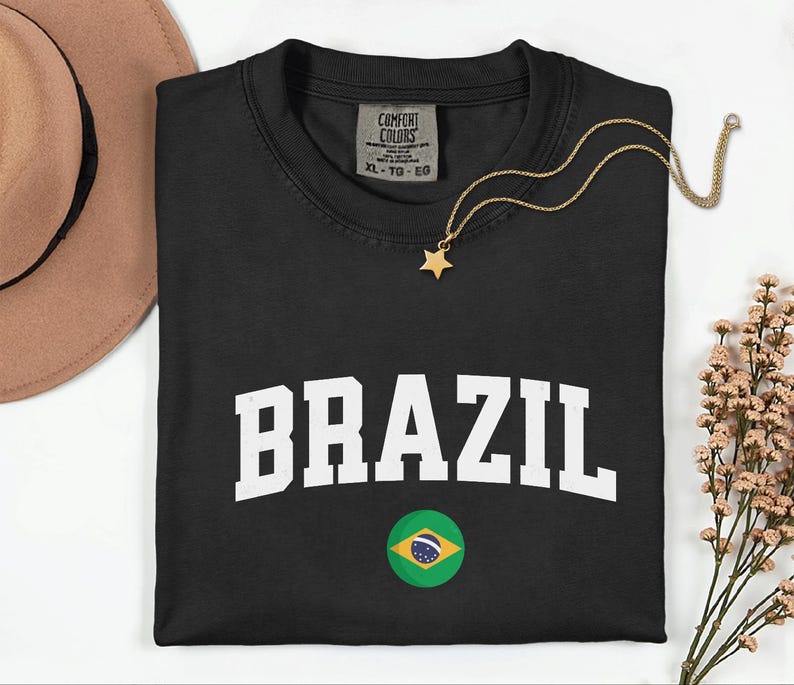 May include: Black t-shirt with the word "BRAZIL" in white block letters and a Brazilian flag graphic. A gold star pendant necklace rests on the shirt. The shirt is a size XL-TG-EG.