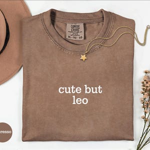 Peut inclure: Un t-shirt &agrave; manches courtes couleur espresso avec le texte blanc "cute but leo" imprim&eacute; sur le devant. Le t-shirt pr&eacute;sente une &eacute;tiquette de marque Comfort Colors &agrave; l'encolure int&eacute;rieure, indiquant une taille XL, et un collier pendentif &eacute;toile.