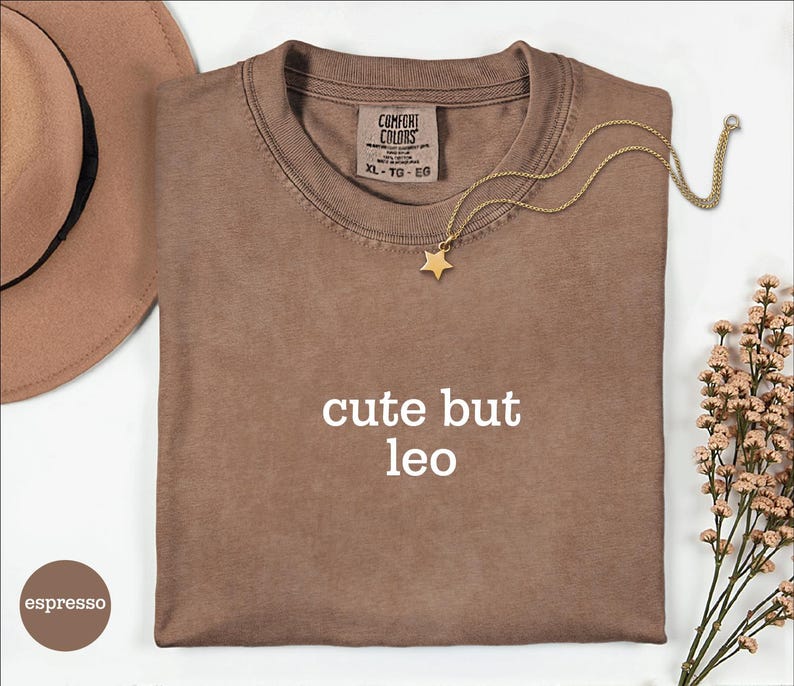 Peut inclure: Un t-shirt &agrave; manches courtes couleur espresso avec le texte blanc "cute but leo" imprim&eacute; sur le devant. Le t-shirt pr&eacute;sente une &eacute;tiquette de marque Comfort Colors &agrave; l'encolure int&eacute;rieure, indiquant une taille XL, et un collier pendentif &eacute;toile.