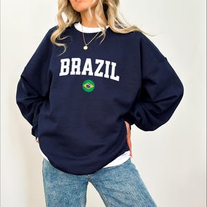 May include: Navy blue sweatshirt with the word "BRAZIL" in white block letters. Below the text is a circular emblem of the Brazilian flag. The sweatshirt has a white crew neck and long sleeves.