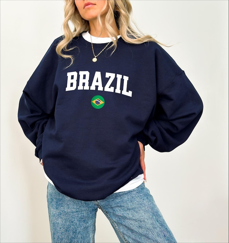 May include: Navy blue sweatshirt with the word "BRAZIL" in white block letters. Below the text is a circular emblem of the Brazilian flag. The sweatshirt has a white crew neck and long sleeves.