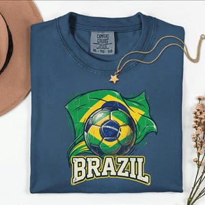 Pode incluir: Uma camiseta azul marinho com uma bola de futebol e a bandeira brasileira em verde e amarelo. A palavra "BRAZIL" est&aacute; impressa em letras brancas abaixo. Um colar de ouro com um pingente de estrela repousa na camiseta.