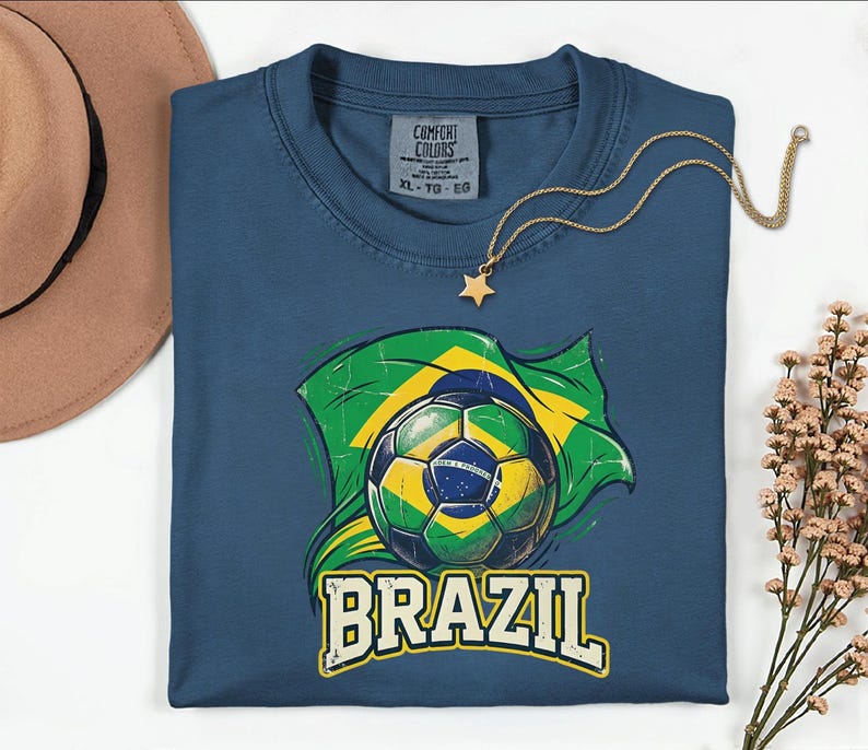 Pode incluir: Uma camiseta azul marinho com uma bola de futebol e a bandeira brasileira em verde e amarelo. A palavra "BRAZIL" est&aacute; impressa em letras brancas abaixo. Um colar de ouro com um pingente de estrela repousa na camiseta.