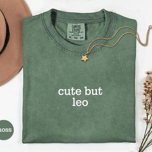 Peut inclure: Un t-shirt vert mousse avec le texte "cute but leo" imprim&eacute; en blanc sur le devant. Le t-shirt pr&eacute;sente une &eacute;tiquette de marque Comfort Colors &agrave; l'encolure int&eacute;rieure, indiquant une taille XL. Un collier dor&eacute; avec un pendentif &eacute;toile est pos&eacute;.