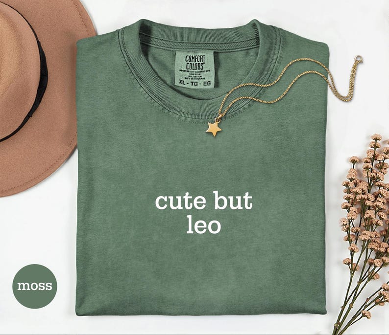 Peut inclure: Un t-shirt vert mousse avec le texte "cute but leo" imprim&eacute; en blanc sur le devant. Le t-shirt pr&eacute;sente une &eacute;tiquette de marque Comfort Colors &agrave; l'encolure int&eacute;rieure, indiquant une taille XL. Un collier dor&eacute; avec un pendentif &eacute;toile est pos&eacute;.