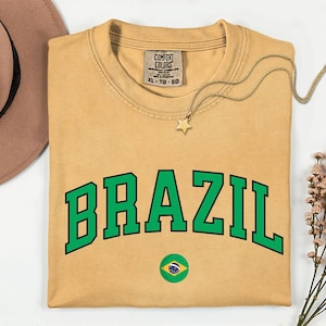 May include: Mustard-colored t-shirt with the word "BRAZIL" in green block letters. A small Brazilian flag is below the text. A gold star pendant necklace and a brown hat are also in the image.