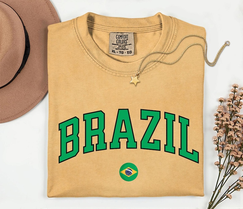May include: Mustard-colored t-shirt with the word "BRAZIL" in green block letters. A small Brazilian flag is below the text. A gold star pendant necklace and a brown hat are also in the image.