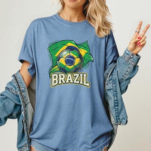 Pode incluir: Camiseta azul clara com um gr&aacute;fico de uma bola de futebol e a bandeira brasileira. A palavra "BRAZIL" &eacute; impressa em amarelo e branco abaixo. A camiseta tem mangas curtas.