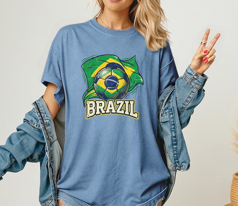 Pode incluir: Camiseta azul clara com um gr&aacute;fico de uma bola de futebol e a bandeira brasileira. A palavra "BRAZIL" &eacute; impressa em amarelo e branco abaixo. A camiseta tem mangas curtas.