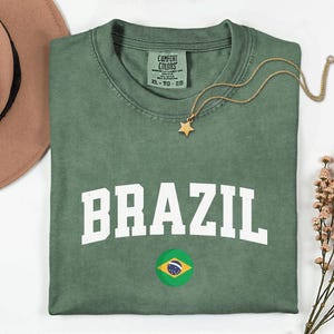 May include: Green t-shirt with the word "BRAZIL" in white block letters and a Brazilian flag graphic. A gold star pendant necklace rests on the shirt. The shirt is size XL.