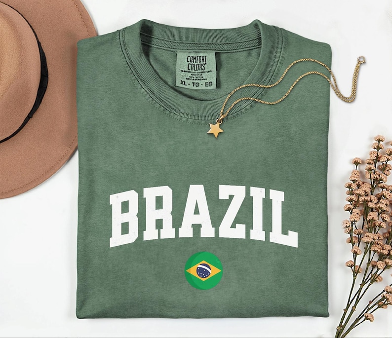 May include: Green t-shirt with the word "BRAZIL" in white block letters and a Brazilian flag graphic. A gold star pendant necklace rests on the shirt. The shirt is size XL.