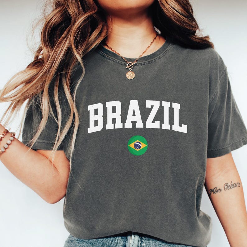 May include: Dark gray t-shirt with the word "BRAZIL" in white block letters and a small Brazilian flag graphic. The shirt is a crew neck style.