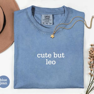 Peut inclure: Un t-shirt bleu jean avec le texte cute but leo imprim&eacute; en blanc sur le devant. Le t-shirt pr&eacute;sente une &eacute;tiquette Comfort Colors au col avec la taille XL-TG-EG. Un collier pendentif &eacute;toile dor&eacute;e repose sur le tissu pr&egrave;s du col.