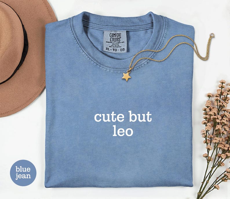 Peut inclure: Un t-shirt bleu jean avec le texte cute but leo imprim&eacute; en blanc sur le devant. Le t-shirt pr&eacute;sente une &eacute;tiquette Comfort Colors au col avec la taille XL-TG-EG. Un collier pendentif &eacute;toile dor&eacute;e repose sur le tissu pr&egrave;s du col.