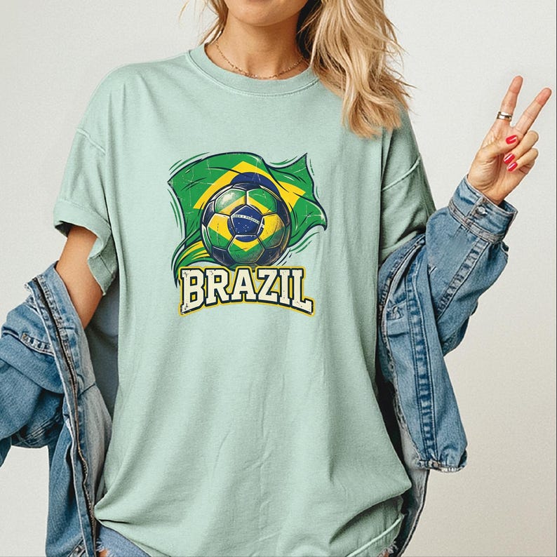 Pode incluir: Camiseta verde claro com uma bola de futebol e a bandeira brasileira. A palavra "BRAZIL" est&aacute; impressa em amarelo abaixo. A camiseta tem mangas curtas e um corte folgado.
