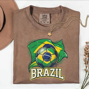 Pode incluir: Uma camiseta marrom com uma bola de futebol e o design da bandeira brasileira. A palavra "BRAZIL" est&aacute; impressa em branco abaixo do gr&aacute;fico. Um colar com pingente de estrela dourada e um chap&eacute;u marrom tamb&eacute;m s&atilde;o vis&iacute;veis. Tamanho XL.