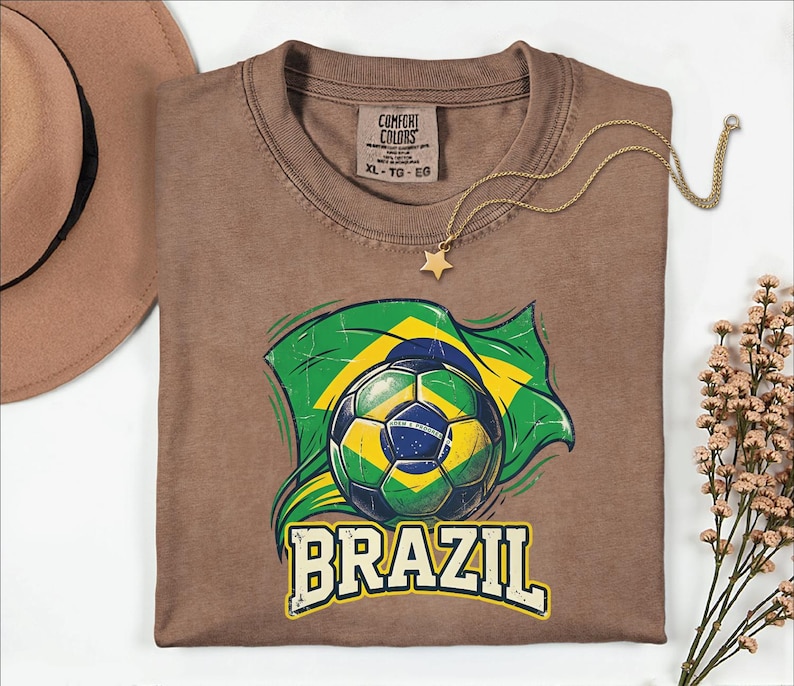Pode incluir: Uma camiseta marrom com uma bola de futebol e o design da bandeira brasileira. A palavra "BRAZIL" est&aacute; impressa em branco abaixo do gr&aacute;fico. Um colar com pingente de estrela dourada e um chap&eacute;u marrom tamb&eacute;m s&atilde;o vis&iacute;veis. Tamanho XL.