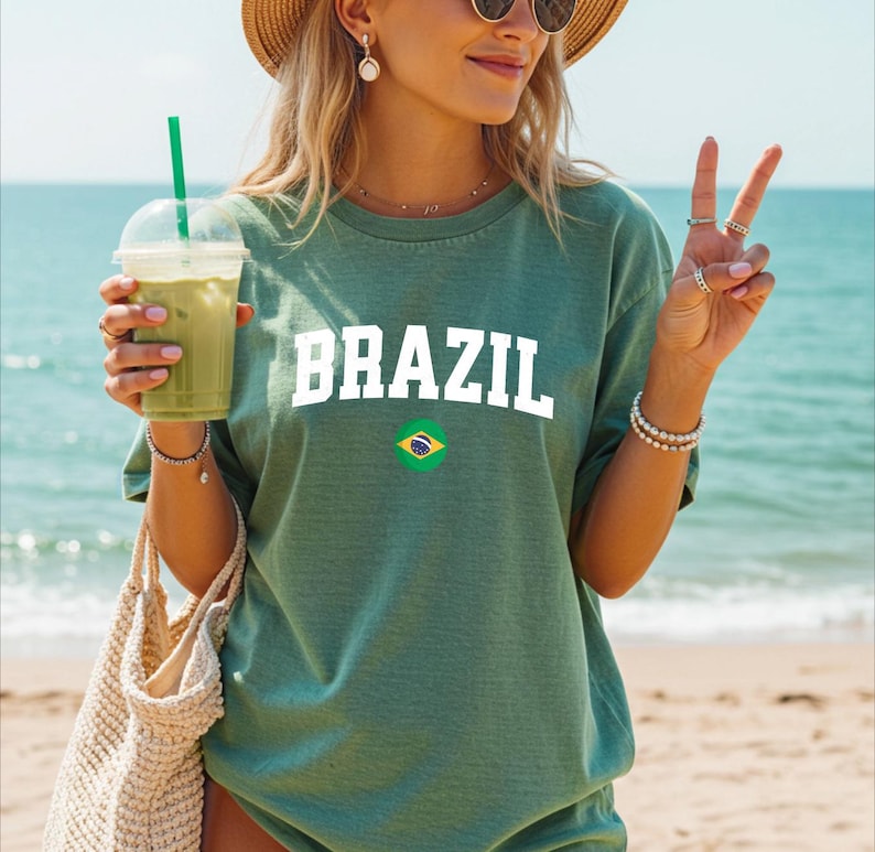 May include: Green t-shirt with the word "BRAZIL" in white block letters and a small Brazilian flag graphic. The shirt is worn on a beach with a drink in hand.